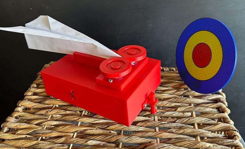 May include: A bright red paper airplane launcher featuring two circular wheels on top and a small trigger mechanism on the side. A white paper airplane is positioned in the launcher, and a circular target with blue, yellow, and red rings stands nearby.