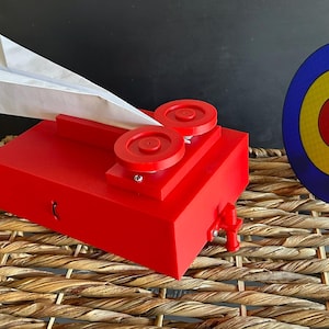 May include: A red paper airplane launcher with red wheels and a white paper airplane. A blue, yellow, and red target is to the right. The objects are on a woven surface.