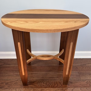 May include: A round side table constructed from light and dark wood. The tabletop features a central dark wood strip flanked by light wood. The four legs are dark wood with light wood inlays, connected by a curved, circular support structure.
