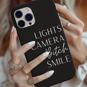 May include: Black phone case with the words "LIGHTS CAMERA Bitch SMILE" in white script. The phone case has a triple-lens camera system. The phone case is being held by a person.