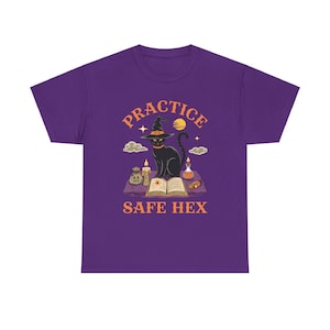 May include: A purple t-shirt featuring a graphic with a black cat wearing a witch hat, sitting on an open book. The text "PRACTICE SAFE HEX" is arched above and below the cat illustration. The design includes a moon, stars, and other witchy elements.