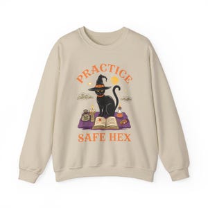Puede incluir: Una sudadera beige con un gato negro que lleva un sombrero de bruja y sostiene un libro. El texto "PRACTICE SAFE HEX" está impreso en la sudadera.