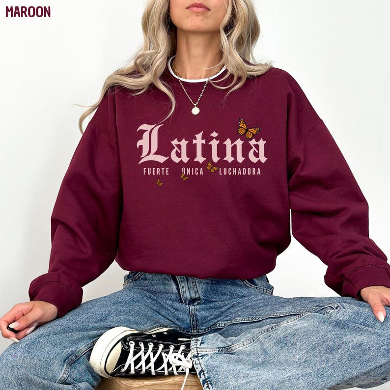 May include: Maroon sweatshirt with the word "Latina" in a decorative font, with "FUERTE UNICA LUCHADORA" below. The design includes butterfly graphics. The sweatshirt is worn with blue jeans and black trainers.