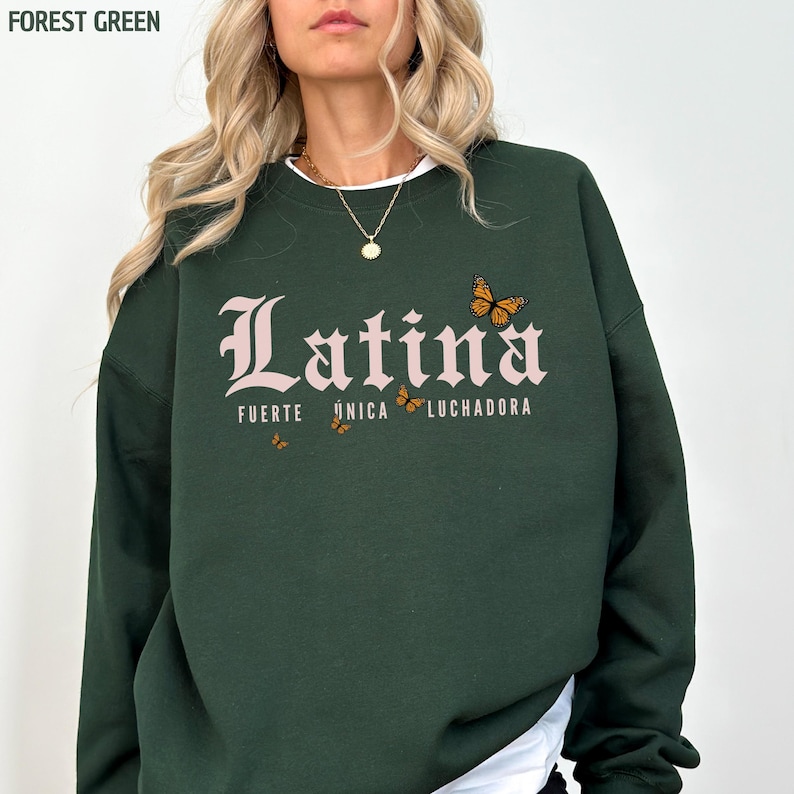 May include: Forest green sweatshirt featuring the word "Latina" in pink script, with "Fuerte, &Uacute;nica, Luchadora" and butterfly graphics. The sweatshirt has a white collar and is made of a soft material.