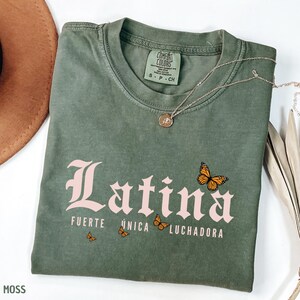 May include: A moss green t-shirt featuring the pink word "Latina", the phrases "Fuerte, &Uacute;nica, Luchadora", and butterfly illustrations. A silver necklace with a pendant is on the shirt.