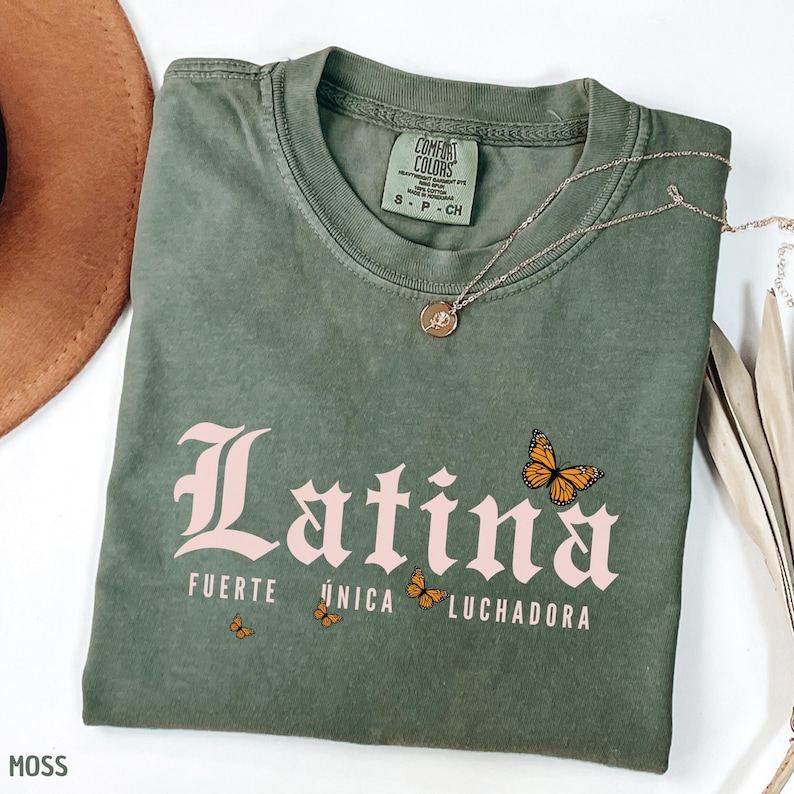 May include: A moss green t-shirt featuring the pink word "Latina", the phrases "Fuerte, &Uacute;nica, Luchadora", and butterfly illustrations. A silver necklace with a pendant is on the shirt.