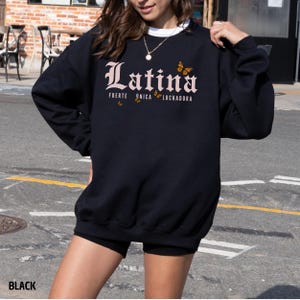 May include: Black crew neck sweatshirt featuring the word "Latina" in a decorative font, with the words "Fuerte, &Uacute;nica, Luchadora" and butterfly graphics. The sweatshirt is a casual style.