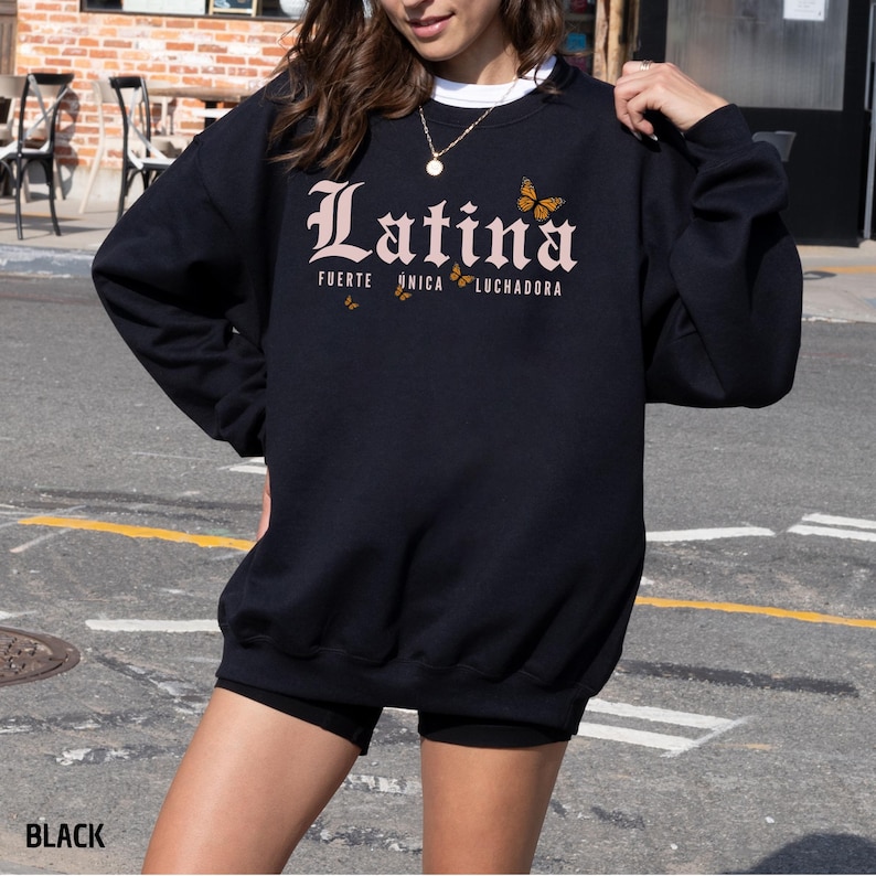 May include: Black crew neck sweatshirt featuring the word "Latina" in a decorative font, with the words "Fuerte, &Uacute;nica, Luchadora" and butterfly graphics. The sweatshirt is a casual style.