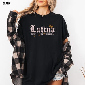 May include: Black crew neck t-shirt featuring the word "Latina" in an ornate font, with the phrases "Fuerte, &Uacute;nica, Luchadora" and butterfly illustrations. The shirt is worn with a plaid flannel shirt.