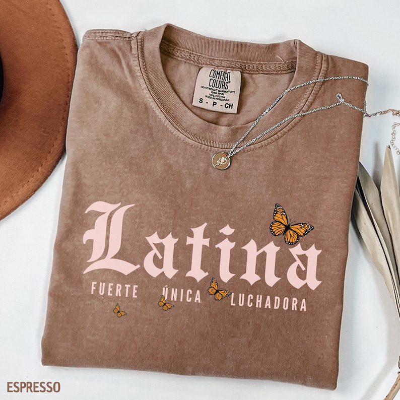 May include: Espresso-coloured t-shirt featuring the word "Latina" in light pink, with "Fuerte, &Uacute;nica, Luchadora" and butterfly graphics. The shirt has a crew neck and short sleeves. A silver necklace with a pendant is draped across the shirt.