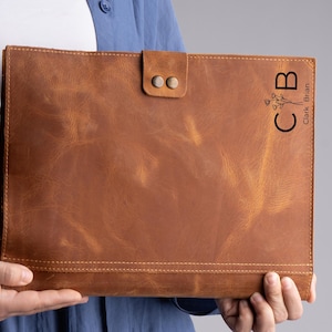May include: A brown leather folder with a flap closure and two metal snaps. The folder has a floral design and the initials "CB" with the name "Clark Brian" printed on the front.