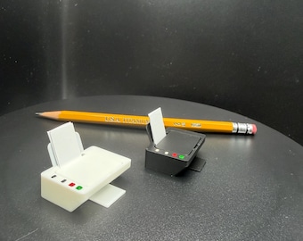 May include: Two miniature printer models, one white and one black, each featuring a small paper tray with white paper inserts and three tiny colored indicator lights on the front panel. A yellow wooden pencil is positioned behind the printers.