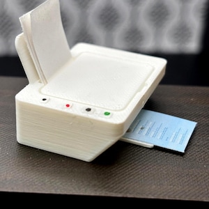 May include: A small, white 3D-printed printer model with a stack of white paper in the input tray. A blue and white card is emerging from the output slot. The printer has red, black, and green buttons on the top.