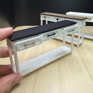 May include: Miniature white console table with a black top and gold drawer pull. The table has a lower shelf and a drawer that is partially open. Two other similar tables are visible in the background, one with a brown top and black drawer pull.