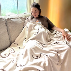 May include: A beige blanket with a repeating pattern of stylized breasts and hearts. The blanket is draped over a person sitting on a beige couch. The person is wearing a brown t-shirt.