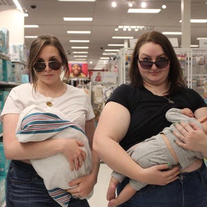 May include: Two people wearing sunglasses stand in a store, each holding a baby wrapped in a blanket. One person wears a white t-shirt and jeans, the other a black t-shirt and jeans. The babies are in gray outfits.