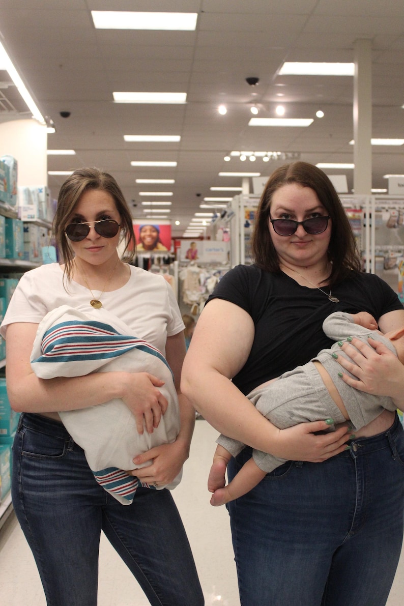 May include: Two people wearing sunglasses stand in a store, each holding a baby wrapped in a blanket. One person wears a white t-shirt and jeans, the other a black t-shirt and jeans. The babies are in gray outfits.