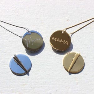 May include: Four necklaces with round pendants. Two pendants say "Mama" in script and block letters. The other two pendants have a bar design. The pendants are gold, silver, and blue. The necklaces are on a white surface.