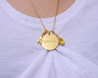 May include: A gold-toned necklace featuring a circular pendant engraved with the word MAMA, accompanied by a letter A charm and a yellow teardrop-shaped crystal charm, all suspended from a thin gold-toned chain against a white fabric background.