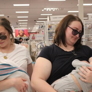 May include: Two women wearing sunglasses are in a store. One is holding a baby wrapped in a blue and white striped blanket. The other is breastfeeding a baby. Shelves of baby products are in the background.