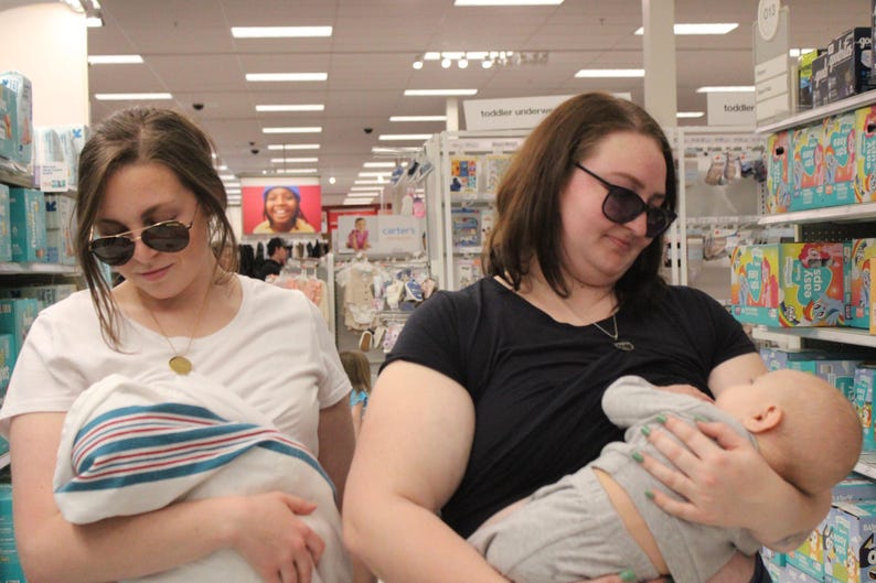 May include: Two women wearing sunglasses are in a store. One is holding a baby wrapped in a blue and white striped blanket. The other is breastfeeding a baby. Shelves of baby products are in the background.