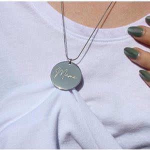 May include: A silver pendant necklace with a round charm. The charm has the word "Mama" engraved in a cursive font. The necklace is displayed on a white t-shirt.