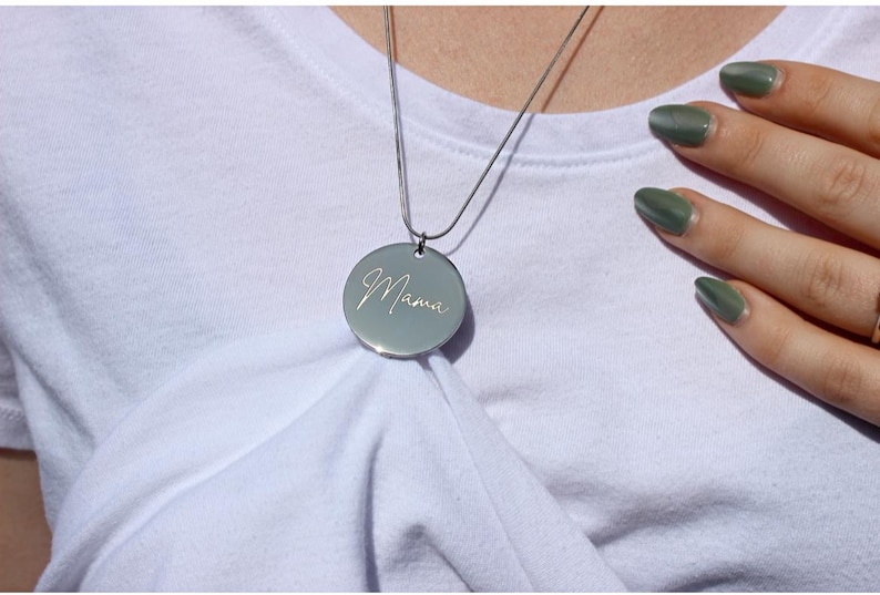 May include: A silver pendant necklace with a round charm. The charm has the word "Mama" engraved in a cursive font. The necklace is displayed on a white t-shirt.