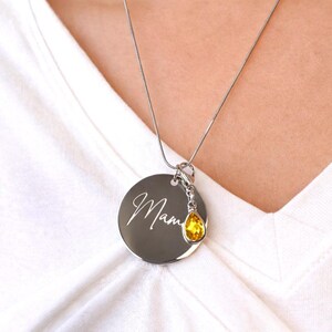 May include: Silver necklace with a round pendant engraved with the word "Mama" in cursive script. A yellow teardrop-shaped charm hangs below the pendant. The necklace is worn over a white shirt.