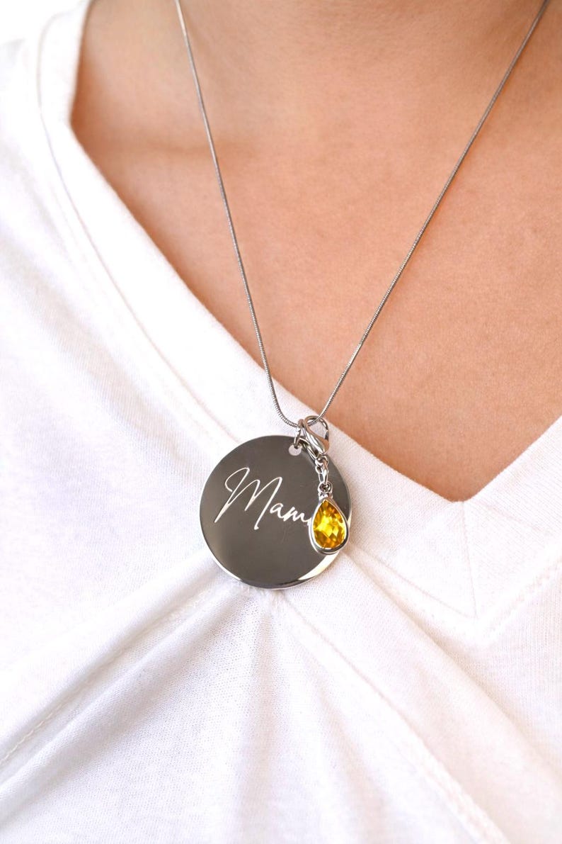 May include: Silver necklace with a round pendant engraved with the word "Mama" in cursive script. A yellow teardrop-shaped charm hangs below the pendant. The necklace is worn over a white shirt.