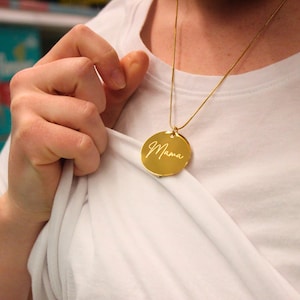 May include: A gold-colored necklace with a round pendant. The pendant has the word "Mama" engraved in a cursive font. The necklace is worn over a white shirt.