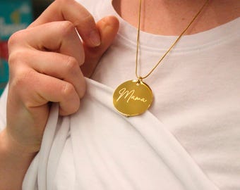 May include: A gold-coloured necklace featuring a circular pendant. The pendant is inscribed with the word "Mama" in cursive script. The necklace is displayed against a white top.