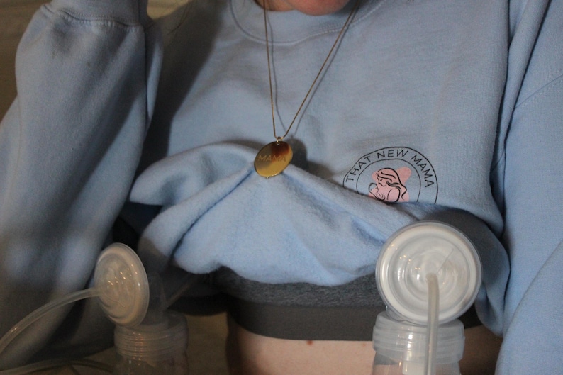 May include: A light blue sweatshirt with the text "THAT NEW MAMA" and a graphic of a mother and baby. A gold pendant hangs from a necklace. Breast pump parts are visible.