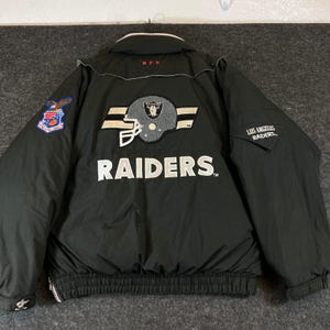 Könnte beinhalten: Eine schwarze Los Angeles Raiders Jacke mit einem großen gestickten Footballhelm-Aufnäher auf der Rückseite, dem Wort RAIDERS in weißer Schrift, einem NFL-Logo am Kragen und einem bunten Adler-Aufnäher auf dem rechten Ärmel mit Los Angeles Raiders Text.