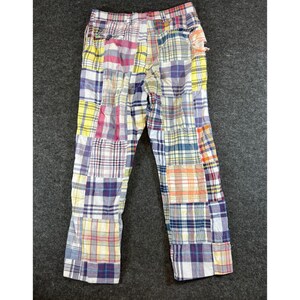May include: Full-length trousers featuring a patchwork design composed of various plaid fabric squares in shades of yellow, blue, red, and white. The pants include a waistband with belt loops, a button closure, and a zippered fly front.