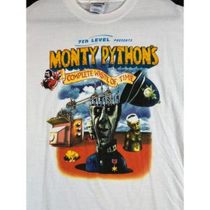 May include: A white t-shirt featuring a graphic print for Monty Python's Complete Waste of Time. The design includes a central figure with an open head, a television set, a police helmet, and various surreal characters against a cloudy sky.