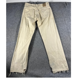 May include: A pair of beige pants featuring a classic five-pocket design with metal rivets, a button fly, and a leather brand patch on the back waistband. The leg hems are frayed and distressed, showing visible wear at the bottom edges.