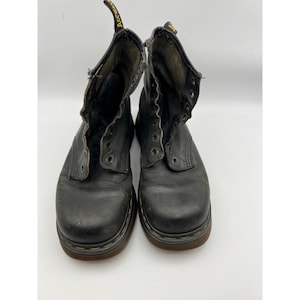 May include: A pair of black leather ankle boots with a rounded toe, featuring yellow stitching along the sole and a black and yellow heel pull tab with the text AirWair. The boots have metal eyelets for laces and a dark brown rubber sole.