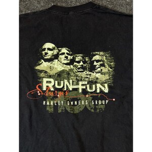 May include: A black t-shirt featuring a graphic of the Mount Rushmore monument in olive green tones. The text reads RUN TO THE FUN, Sturgis, and HARLEY OWNERS GROUP. The design includes a thin orange line tracing a route across the graphic.