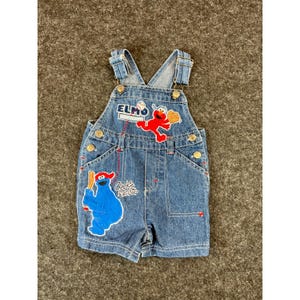 May include: Blue denim overalls featuring embroidered patches of Elmo and Cookie Monster. Elmo is depicted with a baseball glove and the text ELMO, while Cookie Monster holds a bat with the text Cookie Monster. Includes metal hardware.