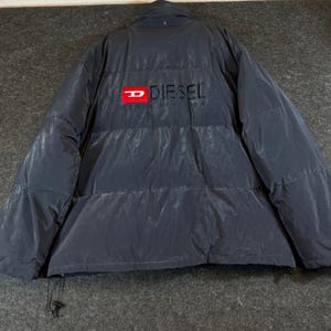 May include: A dark navy blue puffer jacket featuring a high collar and horizontal quilted panels. The back displays a prominent red rectangular logo with the white letter D, followed by the black text DIESEL and DENIM DIVISION below it.