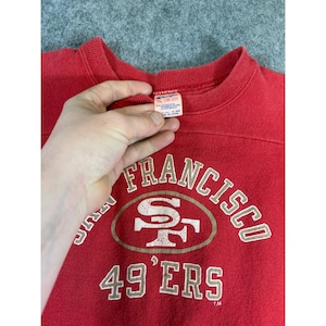 May include: A red crewneck sweatshirt featuring the San Francisco 49ers logo and text in white and gold. The garment has a ribbed collar and a white interior tag indicating size XL (18-20) with fabric composition details clearly visible.