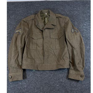 May include: An olive drab wool military jacket featuring a collared neckline, two front chest pockets with buttoned flaps, and long sleeves with buttoned cuffs. Each sleeve displays a chevron rank insignia patch with blue and gold stripes.