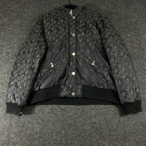 May include: A black quilted jacket featuring a repeating white line-art pattern of abstract figures. The jacket includes a front button closure with silver-toned hardware, a hood, long sleeves with ribbed cuffs, and a wide ribbed hem at the waist.