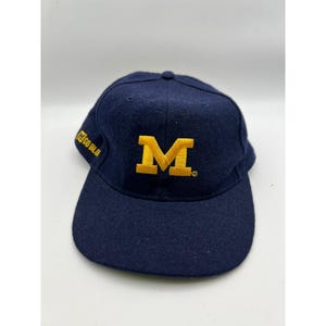 May include: A navy blue baseball cap featuring a large, embroidered yellow letter M on the front centre. The side of the cap displays the embroidered text GO BLUE in yellow. The cap has a structured crown, a curved brim, and a button on top.
