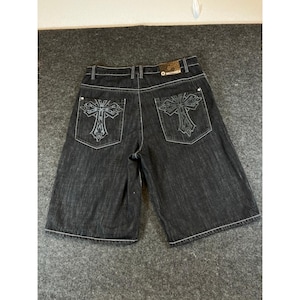 May include: Black denim shorts featuring a prominent cross graphic embroidered in dark grey on each back pocket. The shorts include white contrast stitching, silver-tone metal rivets, and a rectangular brand patch on the back waistband.