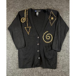 Può includere: Un cardigan nero a maniche lunghe con scollo a V e cinque bottoni frontali. Il capo presenta ricami color oro, tra cui una spirale, una forma a rombo e un motivo a freccia sulla parte anteriore. Due tasche sono posizionate sul fondo.