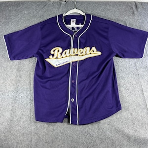 May include: A purple short-sleeved button-down jersey featuring the word Ravens in white and gold script across the chest, with the word Baltimore printed below in smaller text. The garment includes white piping along the collar and front placket.