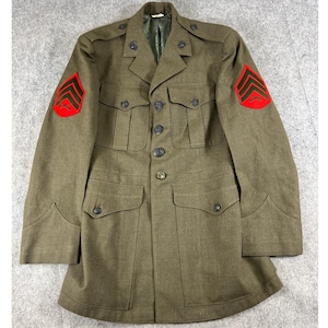 May include: An olive drab military service coat featuring a button-front closure, four exterior pockets with buttoned flaps, and shoulder epaulets. Each sleeve displays a red and black chevron rank insignia patch with a crossed rifle emblem.