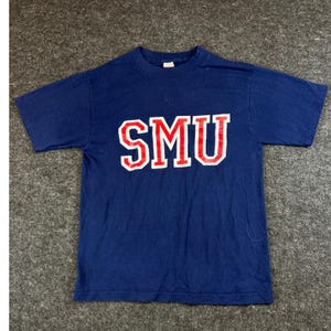 May include: A short-sleeved navy blue t-shirt featuring the letters SMU printed in a bold, block-style font across the chest. The letters are red with a white outline, creating a high-contrast graphic on the solid dark blue fabric of the shirt.