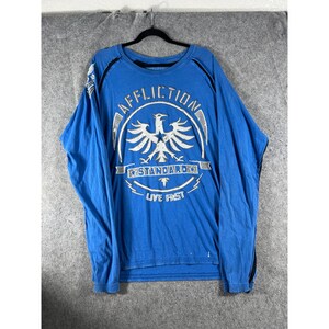 May include: A bright blue long-sleeve shirt featuring a large white graphic on the chest with the text AFFLICTION, STANDARD, and LIVE FAST surrounding a central eagle emblem. The shirt has black trim along the shoulders and sleeves.